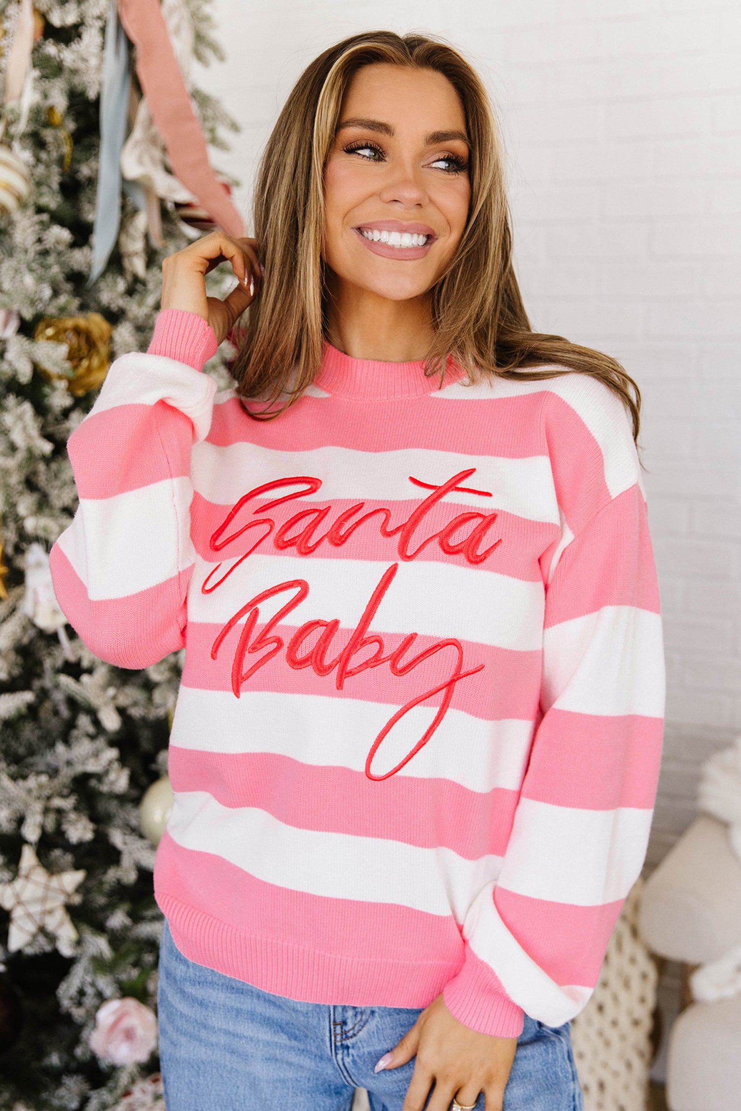 Woman in pink-white striped “Santa Baby” sweater by Christmas tree and white brick wall.