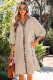 A woman wearing a beige and white vertically striped midi dress with leopard print trim on the collar, placket, and cuffs, holding a brown handbag in an outdoor garden setting.