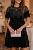 A woman wearing a black velvet short-sleeve dress with sheer mesh sleeves and scalloped floral applique on the bodice, holding a white clutch in a warmly lit indoor setting.