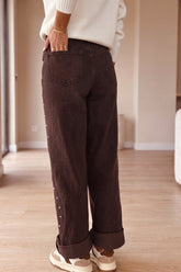 A person shown from the back wearing dark brown wide-leg corduroy pants with rolled cuffs and decorative buttons down the side, paired with a white sweater and white sneakers in a bright indoor room.