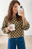 A woman takes a mirror selfie wearing a brown and olive green checkered knit sweater.