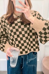 A woman takes a mirror selfie wearing a beige and dark green checkerboard knit sweater, blue jeans.