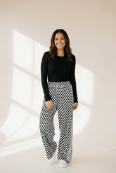 Woman wearing a black top and checkered pants standing against a beige wall.