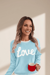 Woman wearing a light blue sweater with 'love' text, holding a red mug against a neutral background