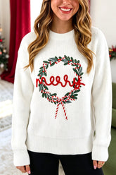 A woman wearing a white ribbed knit sweater with a green and red Christmas wreath and 'Merry' text graphic on the front, posed in a festive indoor setting.