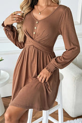 A woman wearing a long-sleeve brown dress with a V-neckline, button details, and a gathered waist, seated on a white stool in a bright indoor setting.