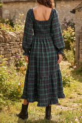 A woman wearing a long-sleeve plaid midi dress with a smocked bodice and tiered skirt, photographed from the back in an outdoor garden setting.