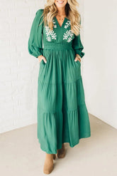 Woman wearing a green dress with floral embroidery against a white brick wall.
