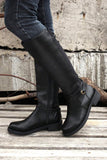 Person wearing black knee-high boots standing on a wooden surface with a concrete wall in the background.