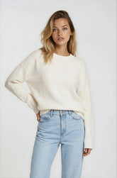 Woman wearing a white sweater and light blue jeans against a plain background