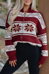 Woman in maroon and cream Nordic snowflake zip-up sweater with black pants by wooden post.