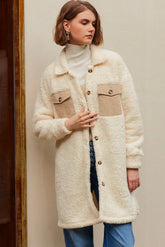 Woman in a cream sherpa coat, white turtleneck, blue jeans, against a beige wall.