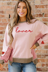 Woman wearing a pink sweatshirt with 'lover' text and heart designs, standing against a brick wall.