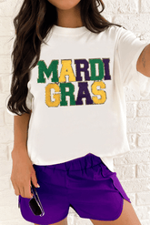 Person wearing a white t-shirt with 'MARDI GRAS' text and purple shorts against a brick wall.