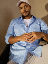 Man wearing a light blue checkered shirt over a white t-shirt, sitting against a neutral background.