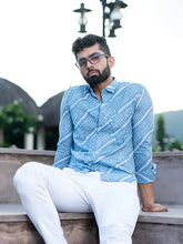 Man wearing a blue patterned shirt and white pants sitting on a ledge with a scenic background.