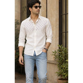 Man wearing a white patterned shirt and light blue jeans standing outdoors.