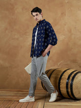 Man wearing a navy patterned shirt and gray pants standing next to a wooden barrel against a brown wall.