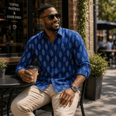 Man in a blue patterned shirt sitting outdoors holding a coffee cup.