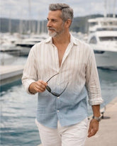 Man wearing a light-colored shirt with a gradient effect, holding sunglasses, by a marina.