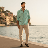 Man in a light green shirt and beige pants standing by a waterfront with buildings in the background.
