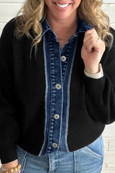 Woman in black ribbed cardigan over a blue denim button-front shirt with light blue jeans against a white brick wall.