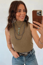 Woman taking a mirror selfie wearing a brown sleeveless top and blue jeans, with layered necklaces and bracelets.