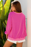Woman wearing a bright pink ribbed sweater and white shorts standing outdoors with plants in the background.