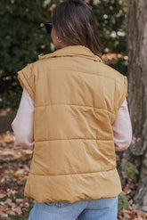 Woman viewed from behind wearing camel tan quilted puffer vest over pink shirt and blue jeans outdoors.