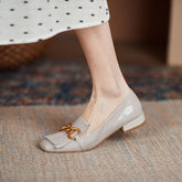 Beige loafers with a gold chain detail worn by a person on a patterned rug.