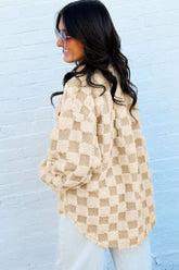 Woman in beige-tan checkered fuzzy sherpa pullover, shown side-back against white brick wall.