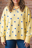 Woman in yellow blue-dot crewneck, blue jeans & beaded necklace by brick wall.