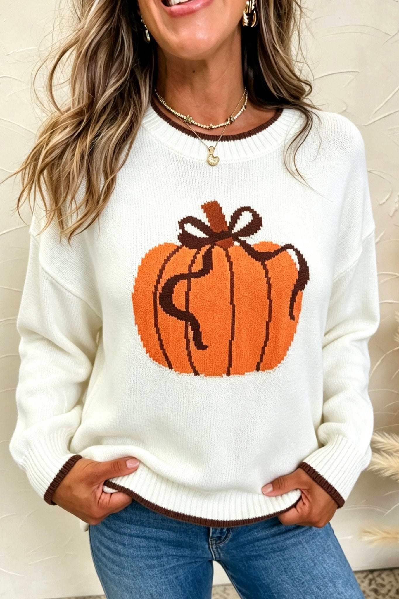 Woman wearing a cream knit sweater with an orange pumpkin graphic and brown trim.