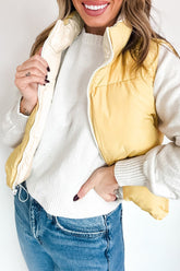 Woman in pale yellow puffer vest over white knit sweater and blue jeans against a light background.