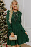 Woman in dark green knit bow-neck dress holding gold patterned clutch by Christmas tree.