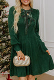 Woman in dark green cable-knit bow-neck dress holding gold clutch by Christmas tree.