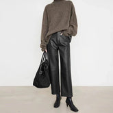 Person wearing a brown sweater and black leather pants holding a black bag against a white background