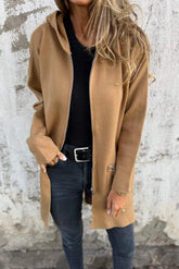 Person wearing a brown coat over a black top and blue jeans against a textured wall.
