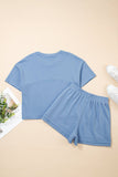 Blue t-shirt and shorts set on a beige surface with a magazine and sneakers in the background.