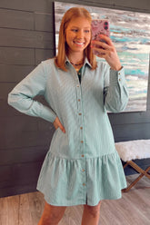 A woman takes a mirror selfie wearing a mint green and white vertically striped long-sleeve button-down shirt dress with a ruffled hem, set against a dark wood-paneled wall.