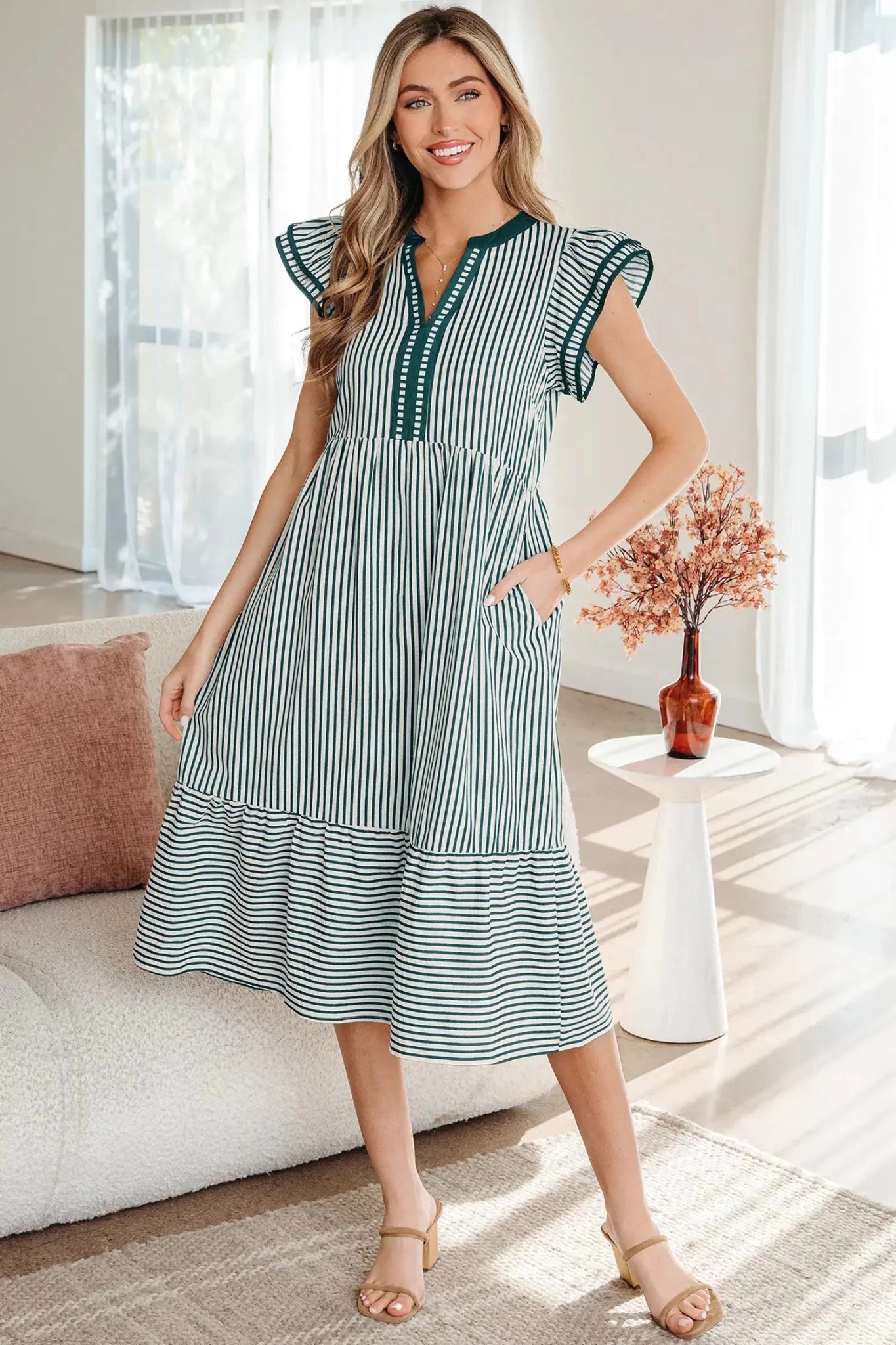 A woman modeling a green and white vertically striped midi dress with ruffled cap sleeves, a split neckline, and a tiered skirt, standing in a bright, modern living room.