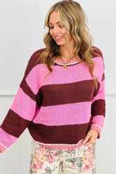 Woman in chunky pink and dark brown striped knit sweater with floral-print pants against a white paneled wall.