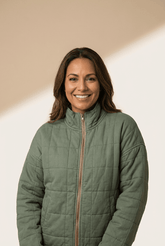 Smiling woman in an olive green quilted zip-up jacket against a warm beige background.