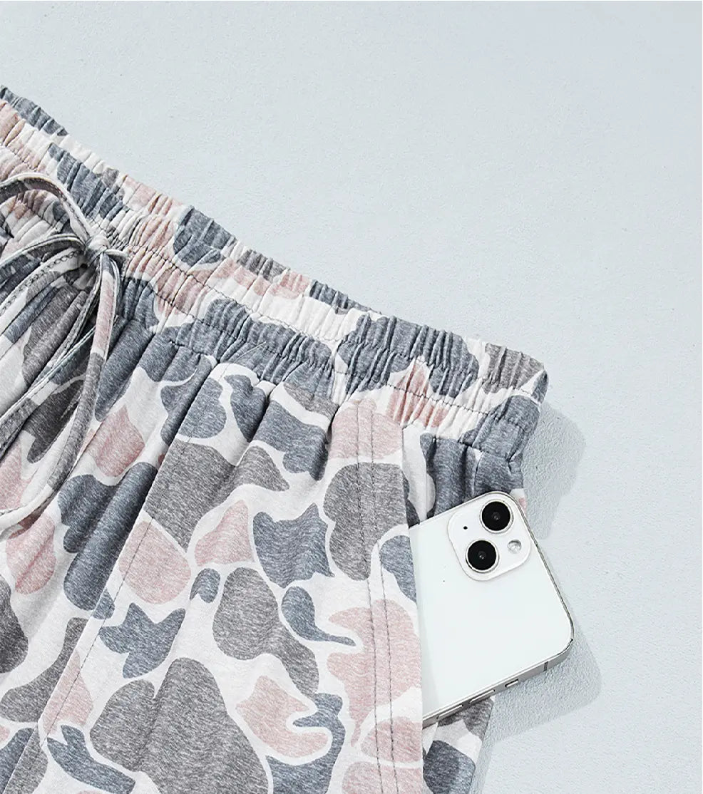 Close-up of a pair of loose shorts with an elastic drawstring waist and a gray, white, and dusty pink abstract print. A white smartphone is tucked into one of the side pockets, set against a light gray background.