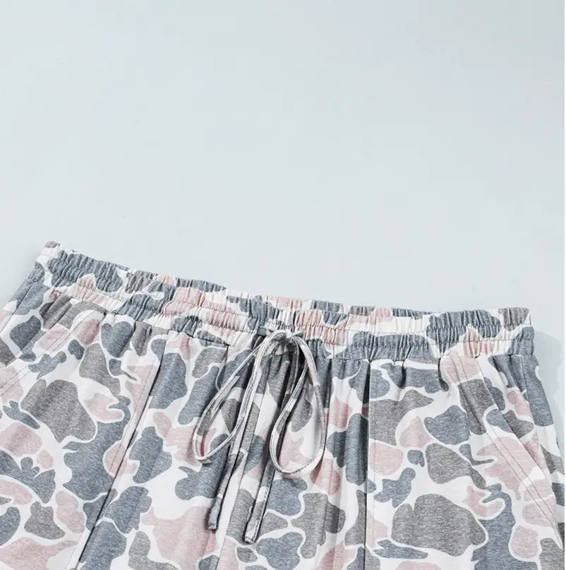 Close-up of a pair of loose shorts with an elastic waistband and drawstring, featuring a gray, pink, and white abstract camouflage pattern, laid flat against a light gray background.