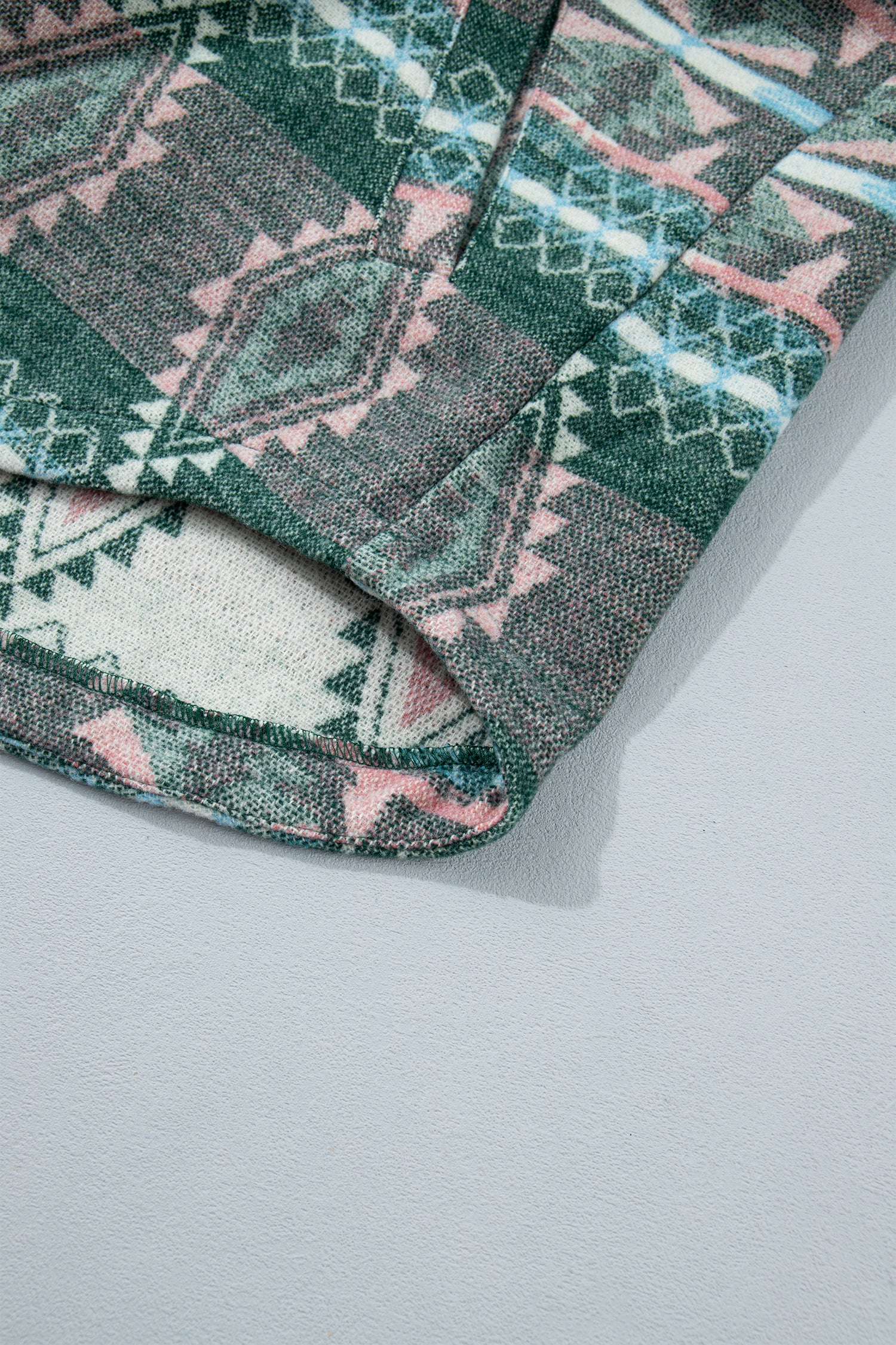 A close-up of a folded garment showing a geometric patterned fabric with shades of green, pink, and off-white on a light gray background.
