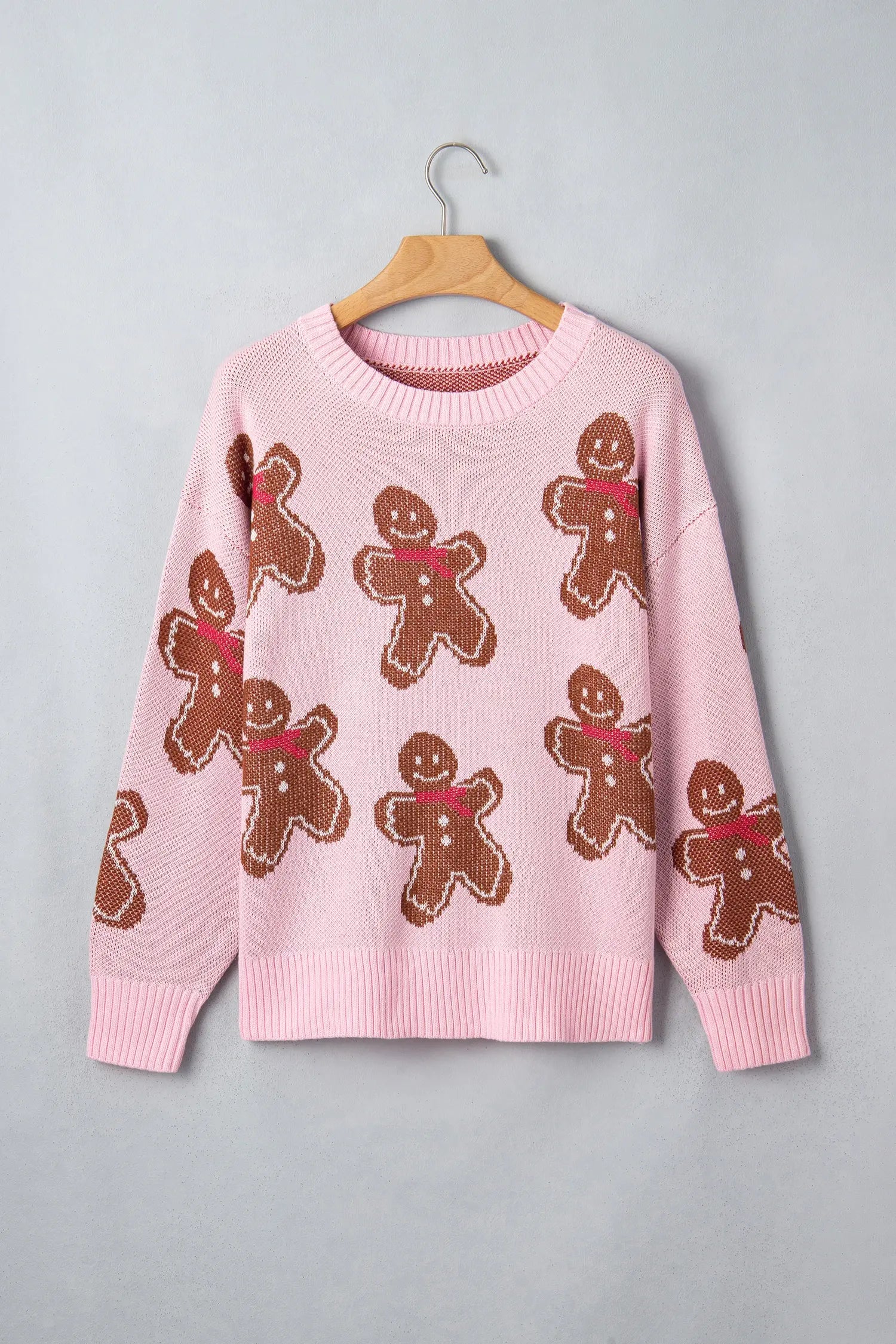Pink crewneck knit sweater with brown gingerbread men in red scarves on hanger against gray.
