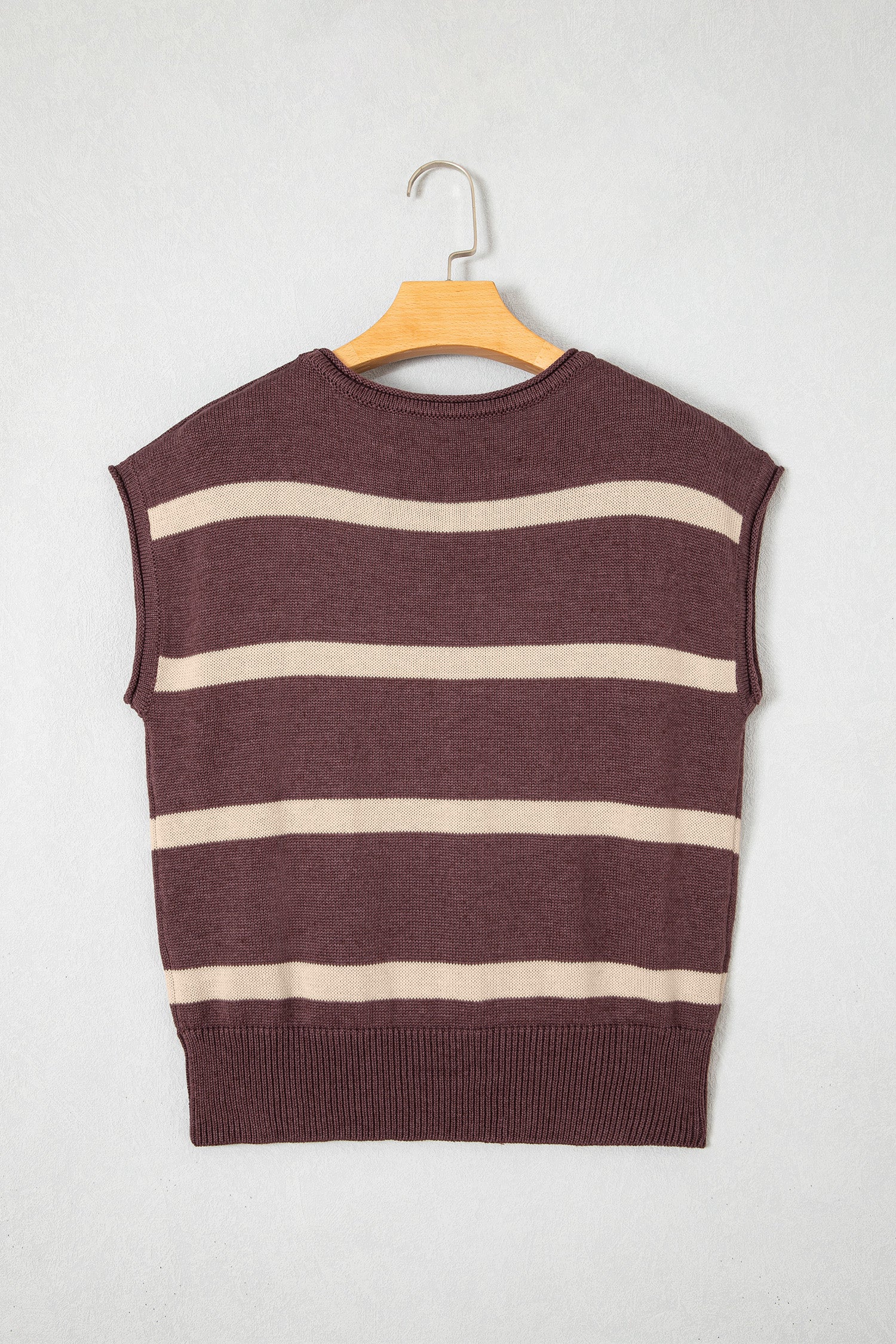 A sleeveless knit sweater vest with horizontal cream-colored stripes on a dark plum-brown base, hanging on a wooden hanger against a light gray textured wall.