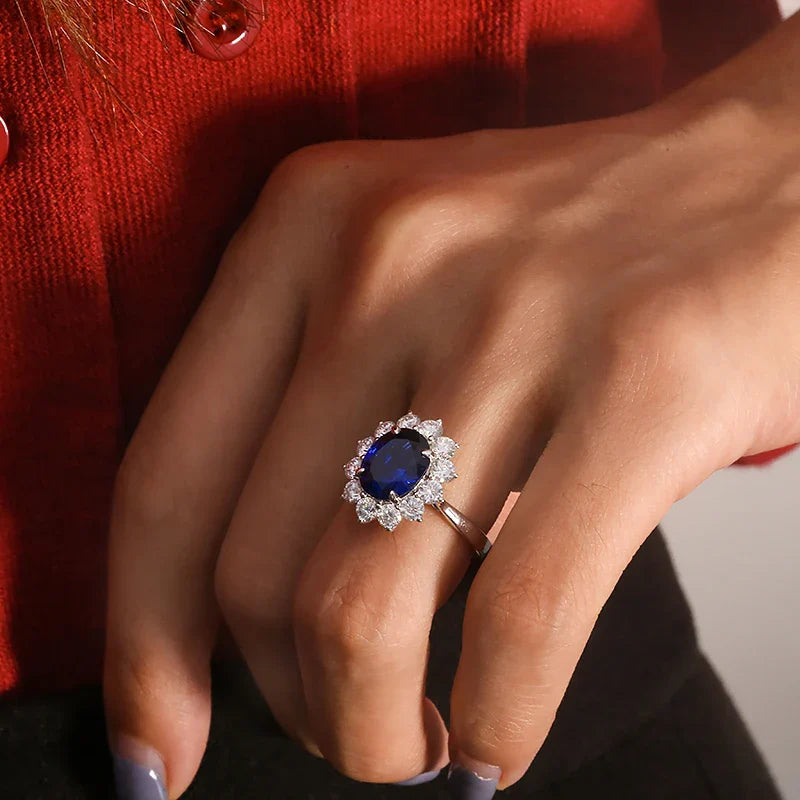 Close-up of a hand wearing a ring with a blue gemstone and diamond halo against a red garment.
