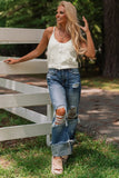 Woman in a white sleeveless top and ripped jeans standing next to a wooden bench outdoors.